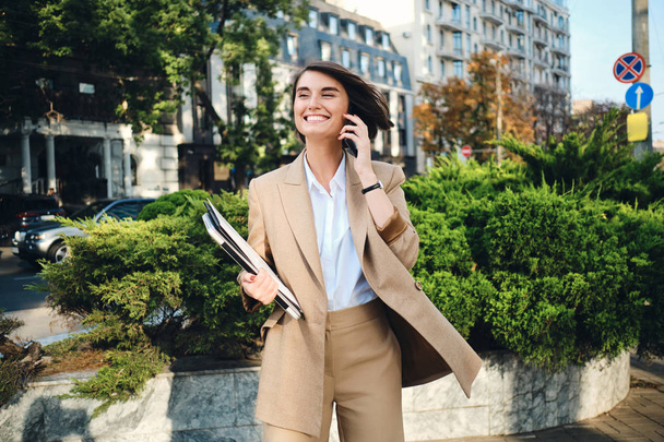 Young cheerful businesswoman in beige suit with laptop happily talking on cellphone on city street - Photo, Image