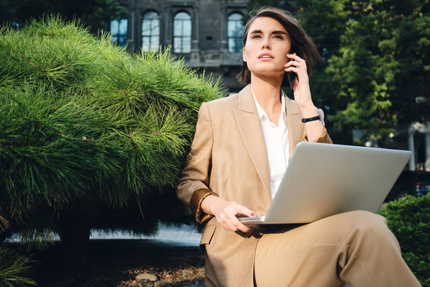 Joven mujer de negocios elegante hablando soñadoramente en el teléfono celular que trabaja en el ordenador portátil al aire libre
 - Foto, Imagen