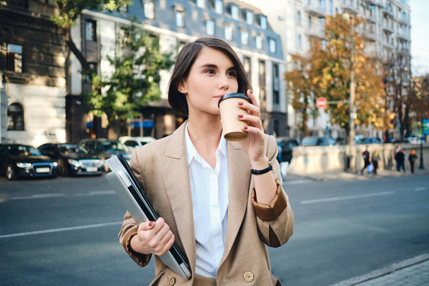 Young pretty businesswoman with laptop drinking coffee during break  on city street - Photo, Image