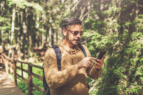 Fashionable man tourist using smartphone while standing on wooden footpath in forest. - Zdjęcie, obraz