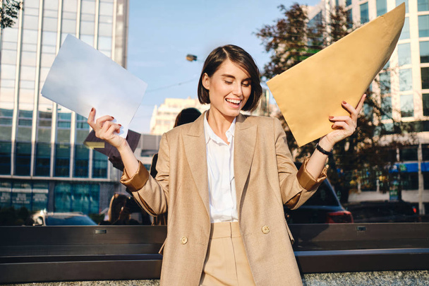 Young cheerful businesswoman holding envelope with response and rejoicing on street - Photo, Image