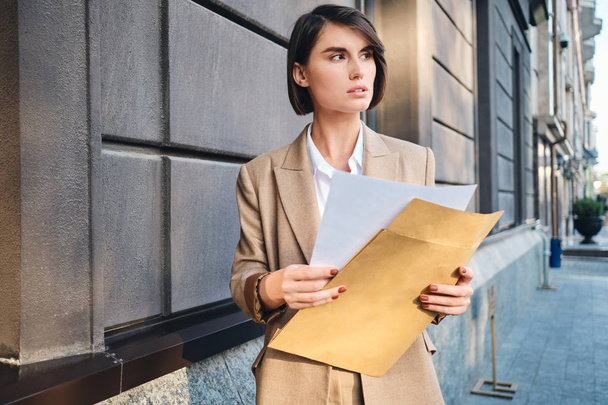 Young serious businesswoman in suit intently looking away working with papers on street - Photo, image