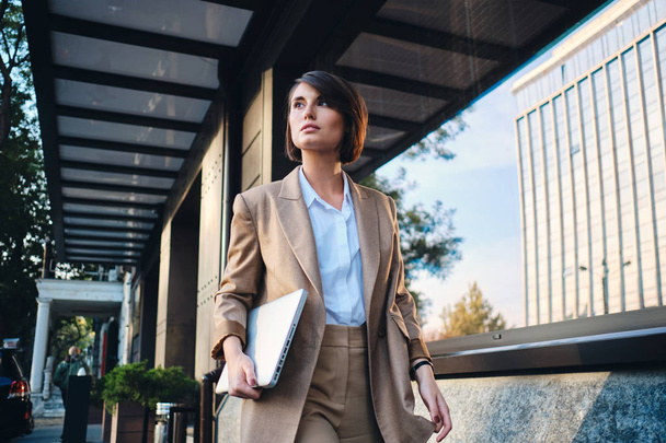Young attractive stylish businesswoman with laptop thoughtfully walking on street - Photo, Image