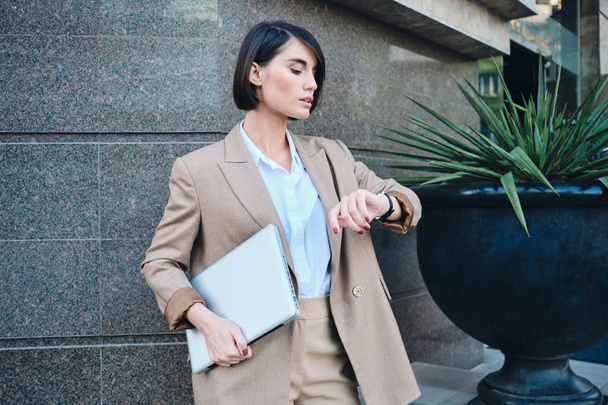 Jovem mulher de negócios elegante confiante em terno bege com laptop assistindo tempo no relógio de pulso ao ar livre
 - Foto, Imagem