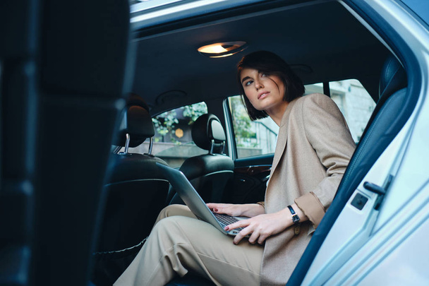 Joven hermosa mujer de negocios con estilo cuidadosamente mirando hacia fuera trabajando en el ordenador portátil en el coche
 - Foto, imagen