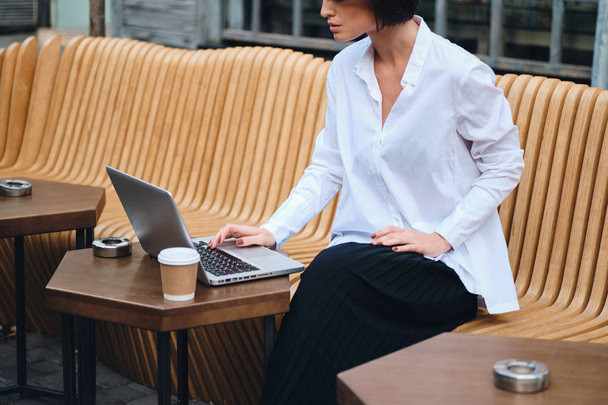 Close up young stylish businesswoman with coffee to go thoughtfully working on laptop in cafe on street - 写真・画像