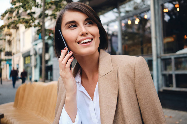 Retrato de jovem empresária alegre alegremente falando no celular no café na rua
 - Foto, Imagem