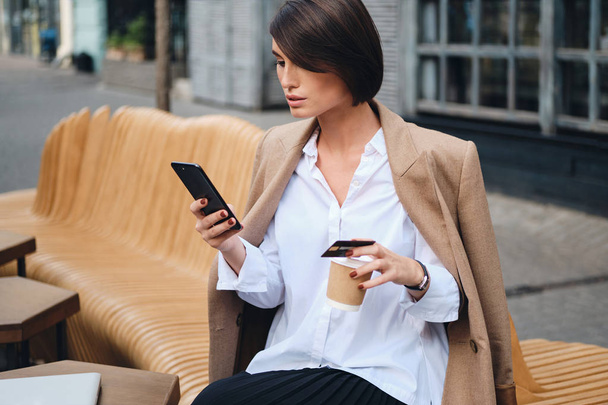 Joven mujer de negocios atractiva con tarjeta de crédito con confianza utilizando el teléfono celular durante el descanso de café en la cafetería en la calle
 - Foto, Imagen
