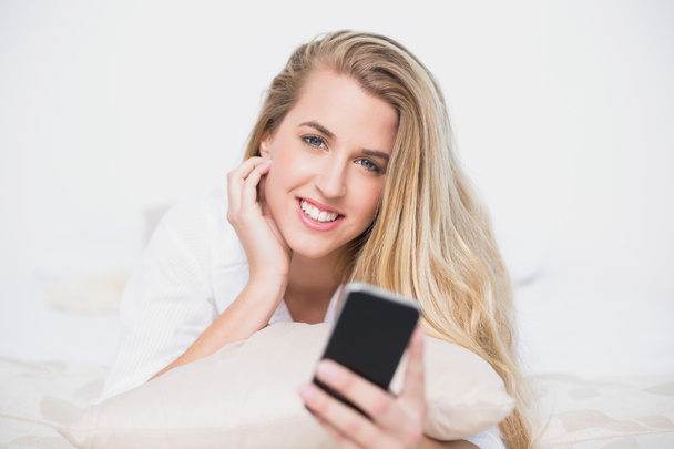 Smiling model holding her smartphone lying on cosy bed - Fotó, kép
