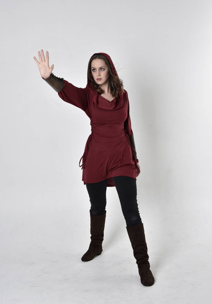 pełnowymiarowy portret brunetki ubranej w czerwoną tunikę fantasy z kapturem. Pozycja stojąca na białym tle studia. - Zdjęcie, obraz