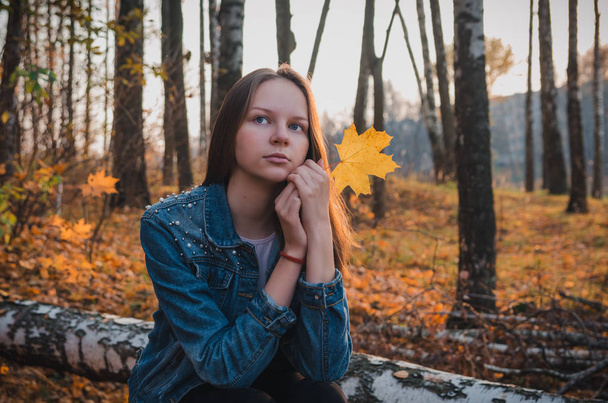 Mooi jong meisje met lang donker blond haar in een blauw denim jasje met een geel esdoorn blad ontspant in het najaarspark in het zonsondergang licht - Foto, afbeelding