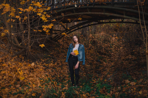 Beautiful young girl with long dark blond hair stands on a path in an autumn park under an old bridge and holds a yellow maple leaf in her hand - Photo, Image