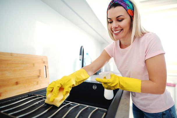 Digne souriante femme blonde caucasienne avec des gants en caoutchouc sur les mains de nettoyage évier de cuisine. Intérieur de cuisine
. - Photo, image