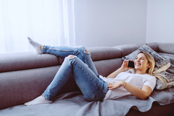 Великолепная улыбающаяся молодая блондинка лежит на диване в гостиной и разговаривает по смартфону. Сплетничать с друзьями
. - Фото, изображение