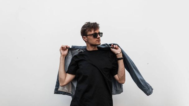 Cool modern young hipster man with a stylish hairstyle in sunglasses in a black T-shirt puts on a vintage blue denim jacket. Attractive urban guy posing near a white wall. Fashionable spring menswear. - Fotografie, Obrázek