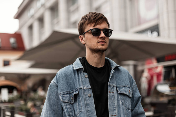 Cool nice young hipster man in a fashionable blue denim jacket in sunglasses stands on the street in the city. Handsome guy model with a stylish hairstyle rests outdoors. Fashion spring menswear. - Fotó, kép