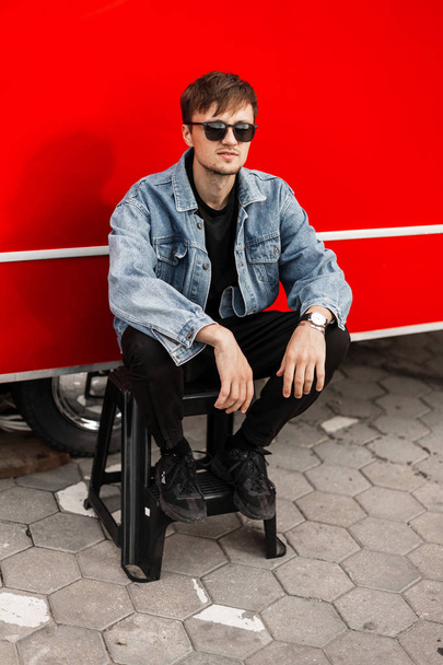 Joven guapo fresco en una chaqueta de mezclilla de moda en pantalones negros de moda en gafas de sol vintage descansando sentado en una escalera cerca de una furgoneta roja moderna en la ciudad. Elegante chico atractivo hipster al aire libre
 - Foto, imagen
