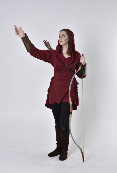 full length portret van een brunette meisje met een rode fantasie tuniek met kap, met een pijl en boog. Staande houding op een witte studio achtergrond. - Foto, afbeelding