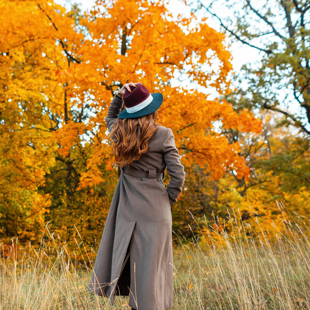 スタイリッシュな秋の服を着たファッショナブルな若い女性が公園の秋の景色を楽しんでいます。シックな帽子のトレンディーなロングコートのエレガントな女の子が森の中に立っています。後ろからの眺め. - 写真・画像