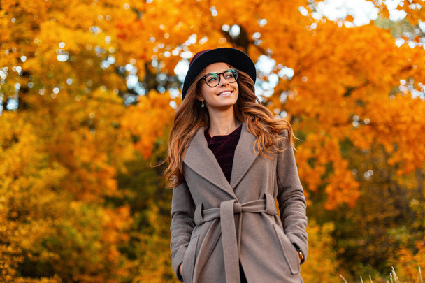 流行のメガネでスタイリッシュなコートでエレガントな帽子に巻き毛で美しい笑顔を持つ肯定的な若い女性は、秋の公園で週末を楽しんでいます。かわいいです幸せなヒップスター女の子ありますリラックス屋外. - 写真・画像