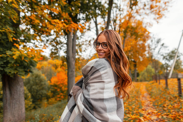 Positive young hipster woman in a fashionable checkered knitted scarf in stylish glasses with a trendy hairstyle walks in the park on an autumn warm day among the trees in yellow-orange foliage.  - Photo, Image