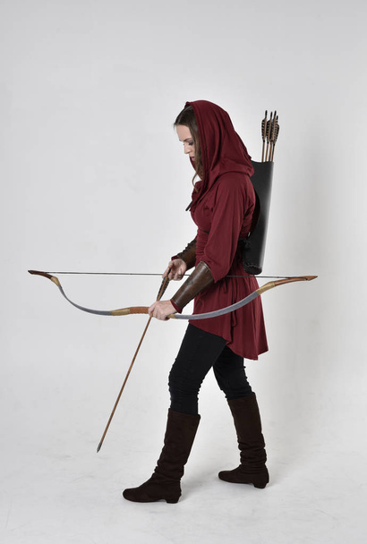 retrato de comprimento total de uma menina morena vestindo uma túnica de fantasia vermelha com capuz, segurando um arco e flecha. De pé pose em um fundo de estúdio branco
. - Foto, Imagem