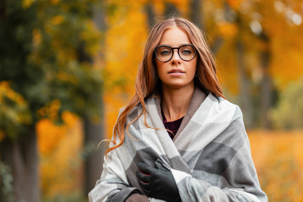 Retrato de uma jovem mulher jovem atraente hipster em óculos elegantes em um casaco em um cachecol de malha vintage em um fundo de folhagem dourada no parque. Menina bonita gosta de um dia quente de outono
 - Foto, Imagem