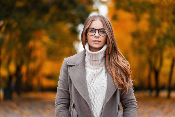Retrato de otoño de una atractiva joven con gafas elegantes en un suéter blanco de moda de punto en un elegante abrigo en un parque sobre un fondo de árboles con hojas de naranja.Chica pasea por el bosque
 - Foto, Imagen