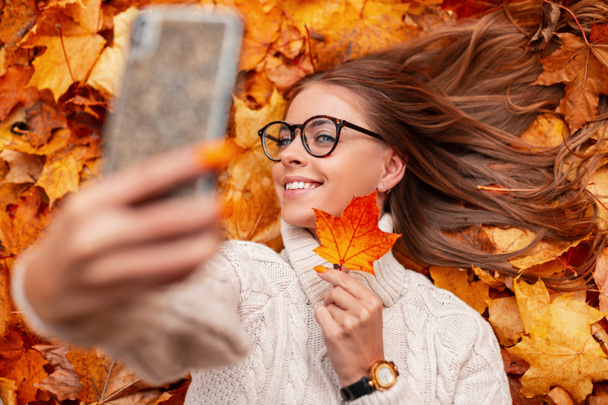 Cheerful young hipster woman in a sweater in a glasses with a beautiful smile holds an orange leaf near face and makes a selfie. Happy girl photographs herself lying in autumn foliage in a park. - Photo, Image