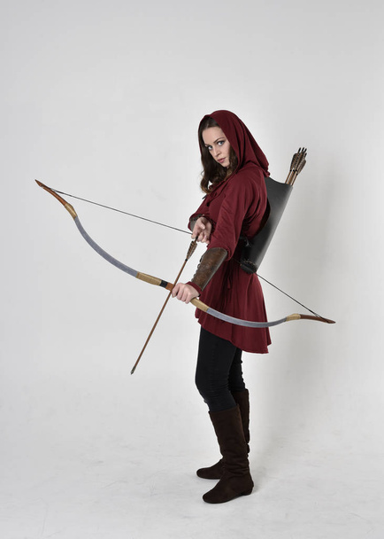 full length portrait of a brunette girl wearing a red fantasy tunic with hood, holding a bow and arrow. Standing pose on a white studio background. - Photo, Image