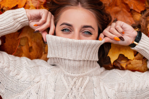 ziemlich modische niedliche junge Frau bedeckt Gesicht mit einem Pullover. attraktives modernes Mädchen mit blauen Augen und natürlichem Make-up liegt auf gelb-orangen Blättern und richtet einen warmen Strickpullover auf. - Foto, Bild