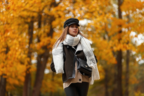 Κομψό νεαρή γυναίκα σε ένα κομψό καπέλο σε ένα κομψό καφέ σακάκι με μια δερμάτινη τσάντα με ένα μαντήλι θέτει στο φόντο των δέντρων με φύλλα χρυσού στο πάρκο σε μια ημέρα του φθινοπώρου. Όμορφο κορίτσι έξω. - Φωτογραφία, εικόνα