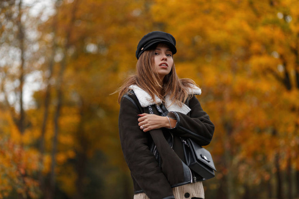 Κομψό νεαρή γυναίκα σε ένα καφέ ζεστό σακάκι σε ένα κομψό καπέλο με ένα γείσο με μια τσάντα vintage δέρμα ποζάρουν στο πάρκο. Μοντέρνο μοντέλο μόδας κορίτσι σε μοντέρνα φθινοπωρινά ρούχα σε εξωτερικούς χώρους. Στυλ νεολαίας. - Φωτογραφία, εικόνα