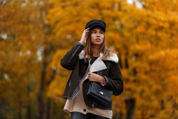 European young woman in stylish clothes in a vintage hat with a black leather bag posing in a park. Attractive trendy girl fashion model in the forest on a background of gold leaves. Autumn style. - Fotó, kép