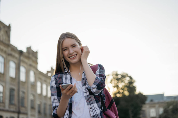 Πορτρέτο της νεαρής ελκυστικής γυναίκας κρατώντας smartphone, ακούγοντας μουσική σε εξωτερικούς χώρους, γέλιο. Ευτυχισμένο hipster κορίτσι χρησιμοποιώντας το κινητό τηλέφωνο, περπατώντας στο δρόμο με σακίδιο πλάτης  - Φωτογραφία, εικόνα