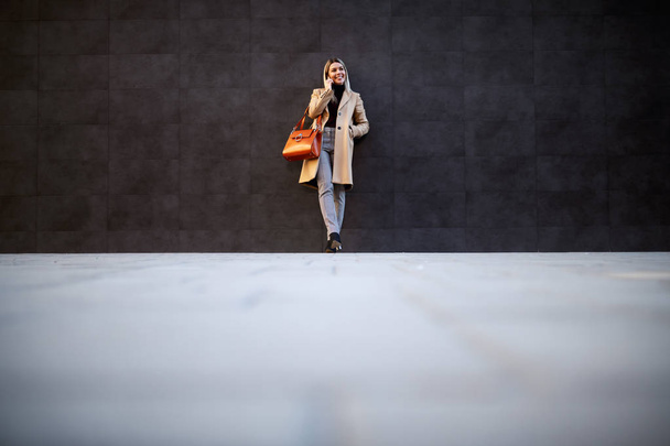 Piena lunghezza di attraente donna bionda caucasica sorridente in cappotto e borsetta appoggiata al muro e parlando sullo smart phone
. - Foto, immagini