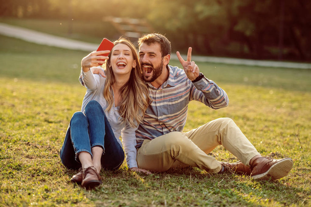 Söpö onnellinen hymyilevä valkoihoinen pari istuu niityllä luonnossa ja ottaa selfie. Mies halaus nainen ja osoittaa voiton merkki, kun nainen tilalla älypuhelin
. - Valokuva, kuva