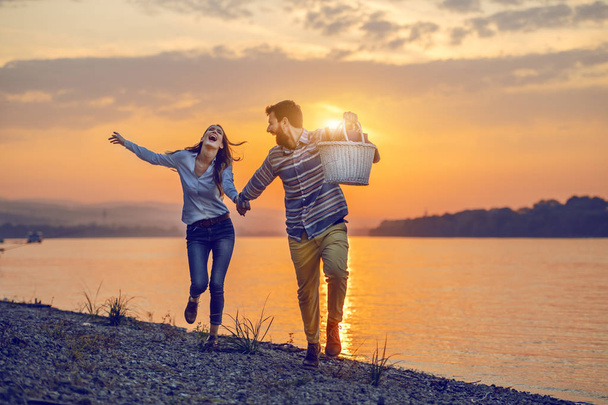 Happy caucasian fashionable couple in love holding hands and running on coast near river. Man holding picnic basket. In background is sunset. - Photo, Image