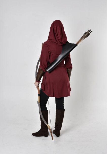 portrét brunetky v červené haleně s kapucí, držící luk a šíp. Stojící póza na bílém pozadí studia. - Fotografie, Obrázek