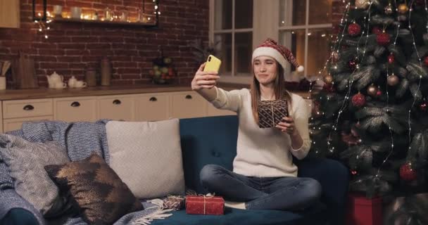 Smiling Woman Wearing Christamas Hat Sitting on the Sofa at Cosy Home Background Holding and Shaking Gift Box near Christmas Tree Making a Video Call or Video Message. Concept of Holidays and New Year - Imágenes, Vídeo