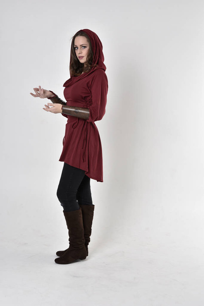 ritratto a figura intera di una ragazza bruna che indossa una tunica fantasy rossa con cappuccio. In piedi posa in profilo laterale su uno sfondo bianco studio
. - Foto, immagini