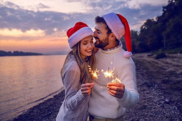 Overjoyed caucasian couple in love standing on coast near river and holding sparklers. Both are dressed in white sweaters and having santa hats on heads. - Valokuva, kuva