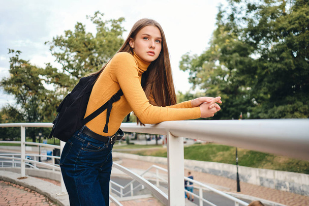 Beautiful casual student girl with backpack intently looking away in city park - Фото, зображення