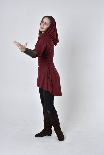 pełnowymiarowy portret brunetki ubranej w czerwoną tunikę fantasy z kapturem. Pozycja stojąca w bocznym profilu na białym tle studia. - Zdjęcie, obraz