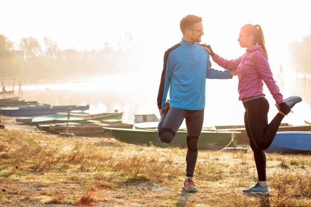 Piękna szczęśliwa młoda para rozciągająca się nad jeziorem wczesnym rankiem, rozgrzewająca się do joggingu - Zdjęcie, obraz