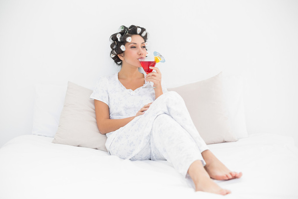 Smiling brunette in hair curlers sitting on her bed drinking a cocktail - 写真・画像