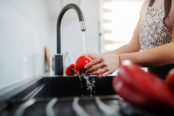 Blisko kaukaska godna kobieta w fartuchu myjąca czerwony pieprz w zlewie kuchennym. Koncepcja przygotowania lunchu. - Zdjęcie, obraz