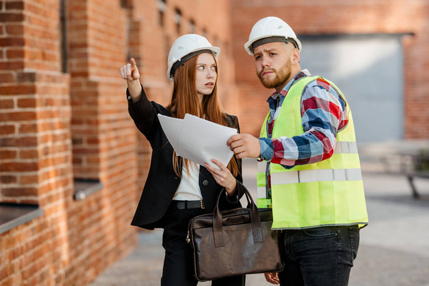 Engineer in hard hat and vest in advance to discuss the project with the architect girl on brick wall background - Photo, Image