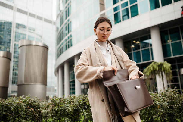 A beautiful Asian looking lawyer girl stands in the background of the business district and looks for a work contract in her leather briefcase - Foto, afbeelding