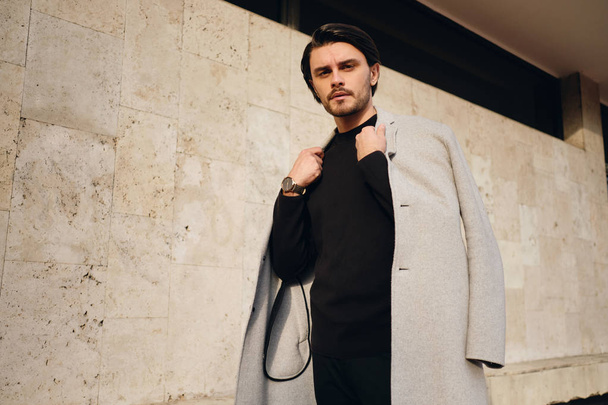 Young serious stylish brunette man with coat on shoulders thoughtfully looking in camera outdoor - Photo, image